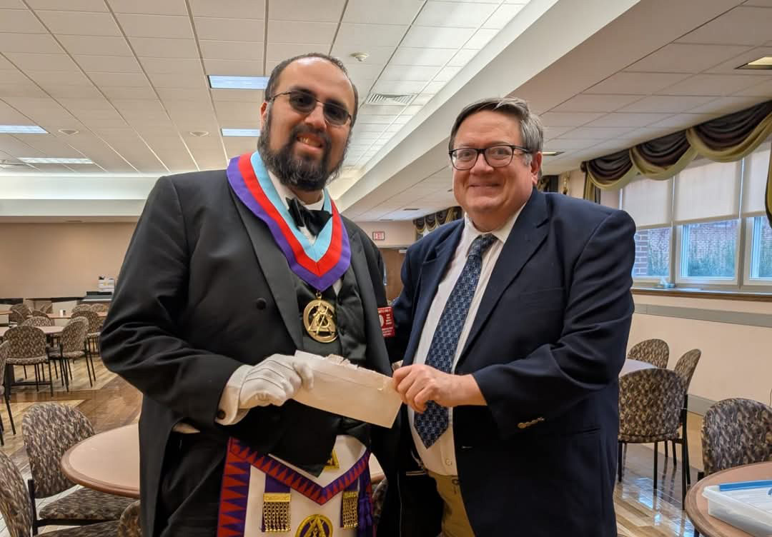 Two men, one dressed in Masonic attire and one dressed in jacket and tie holding check together.